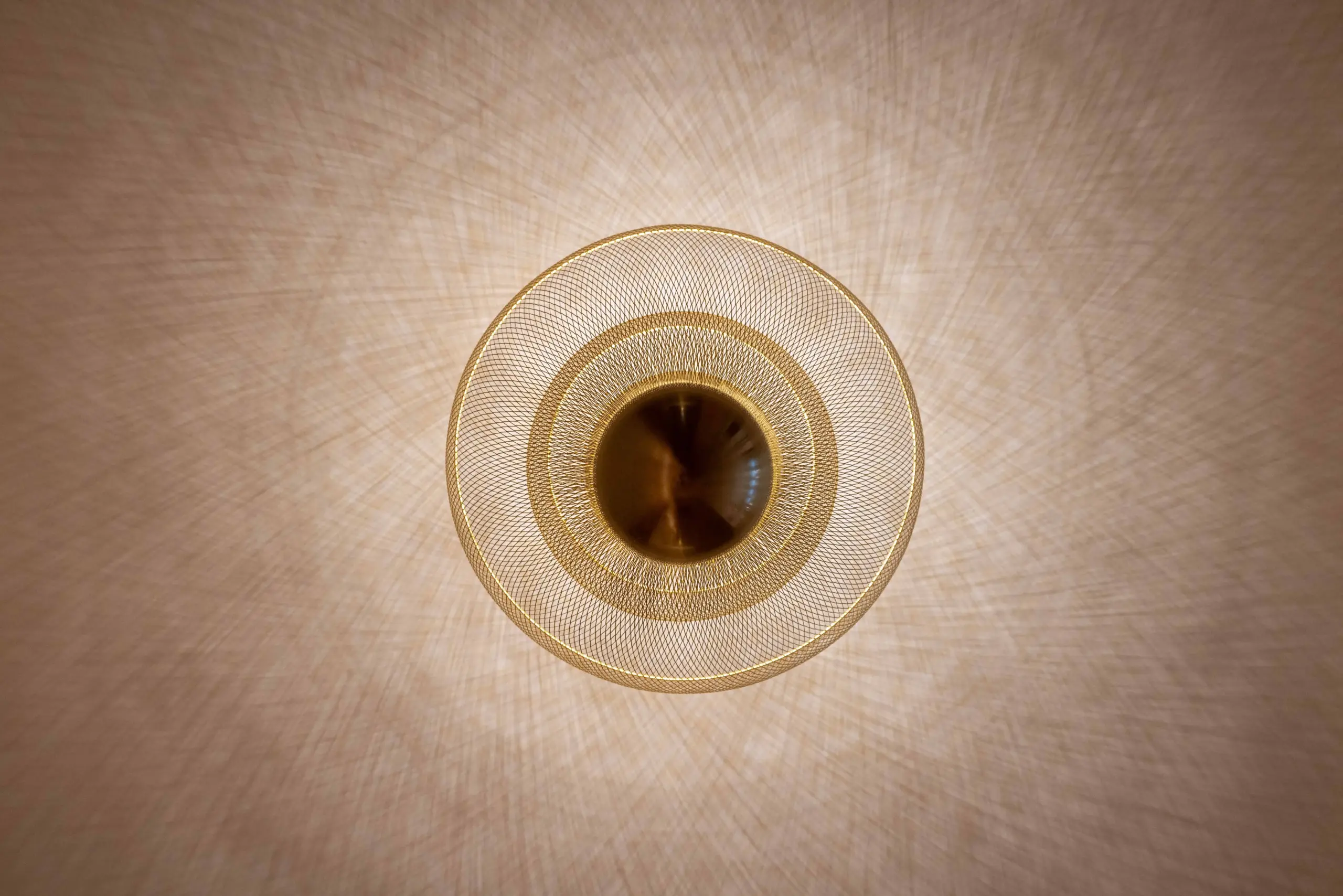 Overhead view of a modern circular ceiling light at Mosque Twenty-Seven, made of layered gold mesh rings against a light textured ceiling