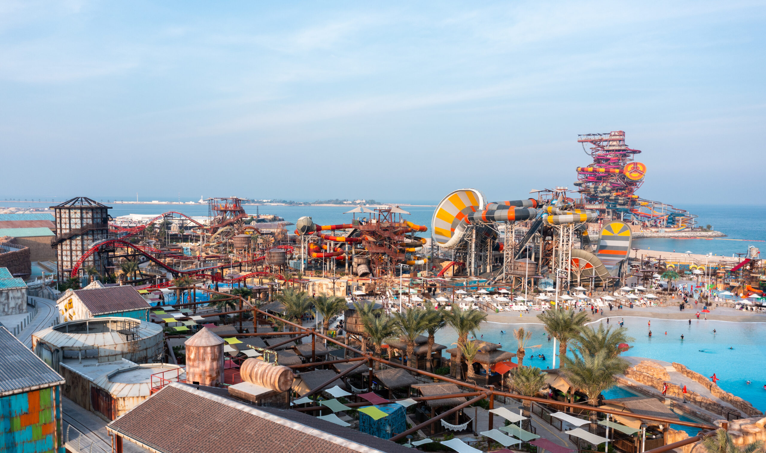 A mid-level photograph of the Meryal Waterpark, showing a multitude of colorful water slides, pools, palm trees, and the industrial-themed RIG 1938 tower in the distance.