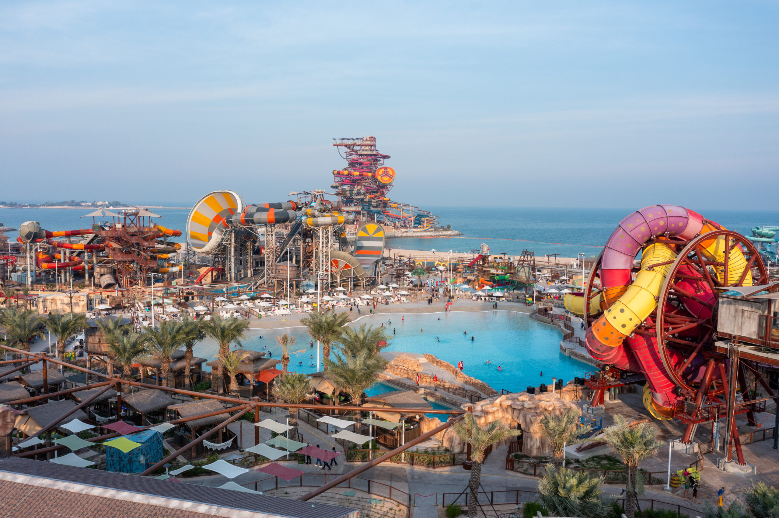 A high-angle view of a wave pool and leisure area within the Meryal Waterpark, showing a rustic-themed pool bar and a red roller coaster track looping over the water.