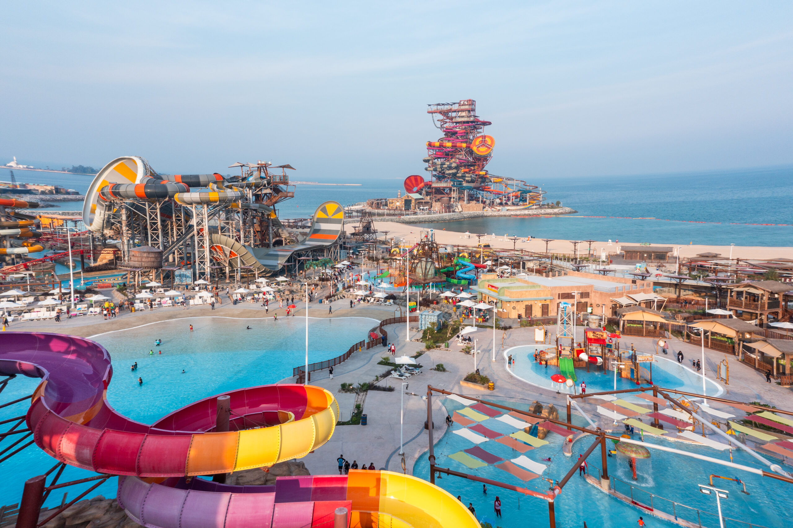 A high-angle view of a wave pool and leisure area within the Meryal Waterpark, showing a rustic-themed pool bar and a red roller coaster track looping over the water.